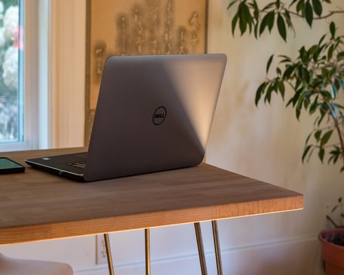 A modern laptop sitting on a clean wooden desk near a window with soft natural sunlight streaming inside